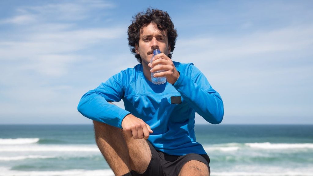 A man in blue sportswear drinks water while sitting by the ocean, enjoying a sunny day.